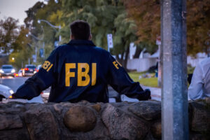 Male FBI agent wearing dark blue coat with FBI logo looking down the street with cars in the dusk seen from behind.